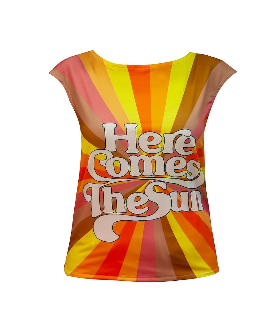 Fotografía de una camiseta con un texto en primer plano que dice lo siguiente: "Here comes the sun" y en el fondo, diferentes rayas, todas del mismo grosor, pero de diferentes colores y todas van al centro.