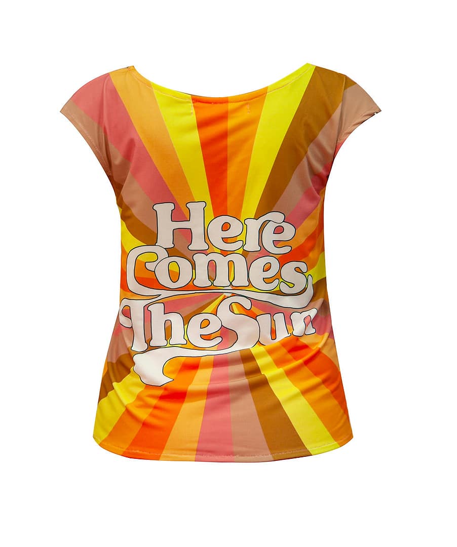 Fotografía de una camiseta con un texto en primer plano que dice lo siguiente: "Here comes the sun" y en el fondo, diferentes rayas, todas del mismo grosor, pero de diferentes colores y todas van al centro.